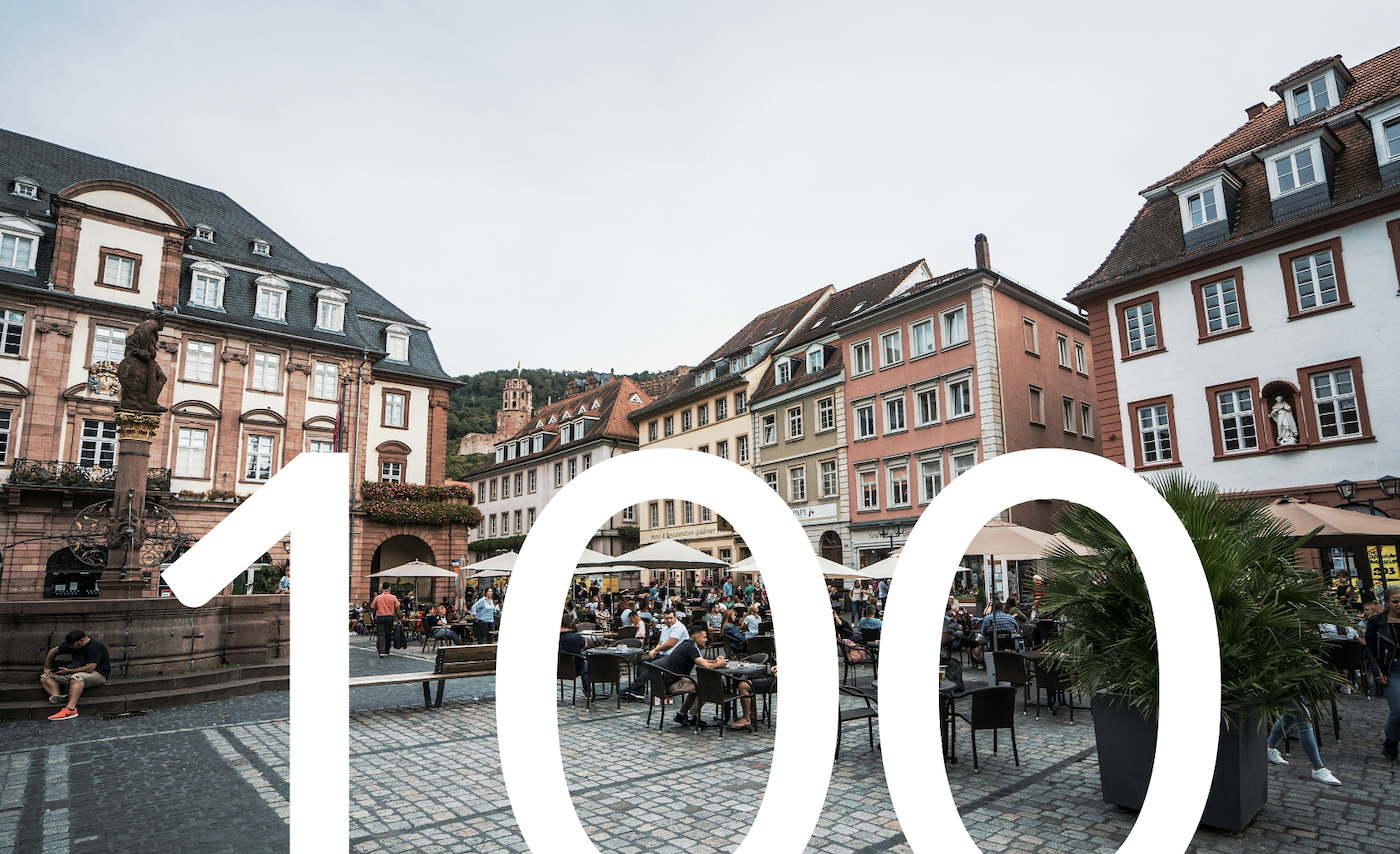 Menschen sitzen auf dem Marktplatz in Heidelberg, auf dem Bild steht groß die Zahl 100